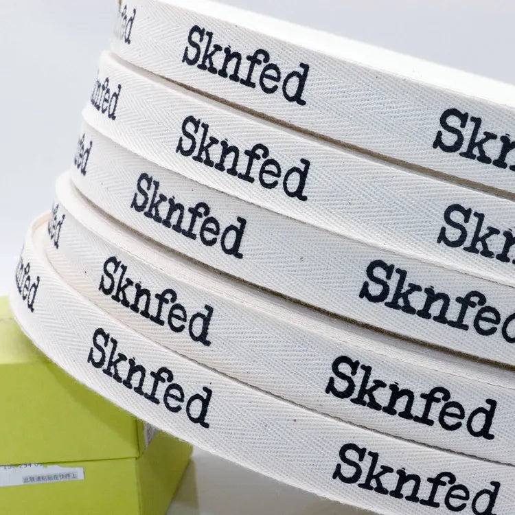 Stacked rolls of natural white herringbone cotton tape with printed black brand names