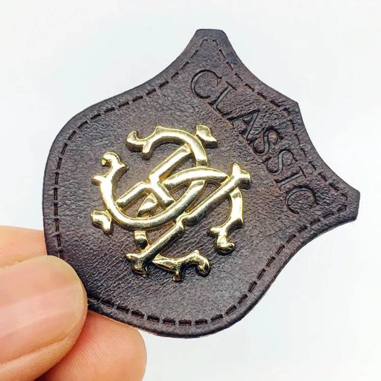 Shield-shaped dark brown leather patch with antique gold metal monogram crest, debossed Classic lettering, and stitched edge for jackets and caps