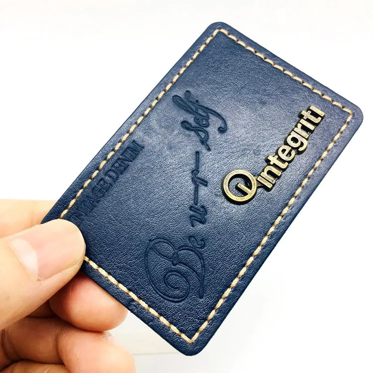 Navy blue custom leather patch featuring antique brass metal logo lettering, debossed script, and contrast perimeter stitching for denim jeans