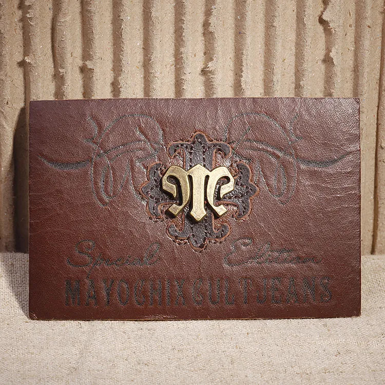 Distressed dark brown leather patch featuring ornate tooled scrollwork, antique brass metal emblem, and debossed denim script