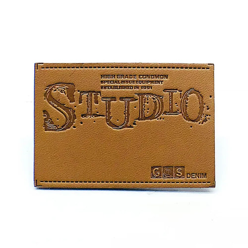 Tan leather patch with debossed vintage Studio wordmark, denim specifications, and precision perimeter stitching for jeans waistband