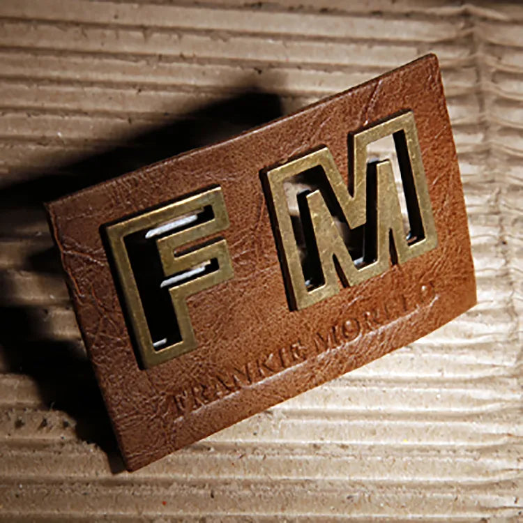 Cognac saddle brown leather patch with raised cutout antique brass metal letters FM and debossed Frankie Morello brand lettering