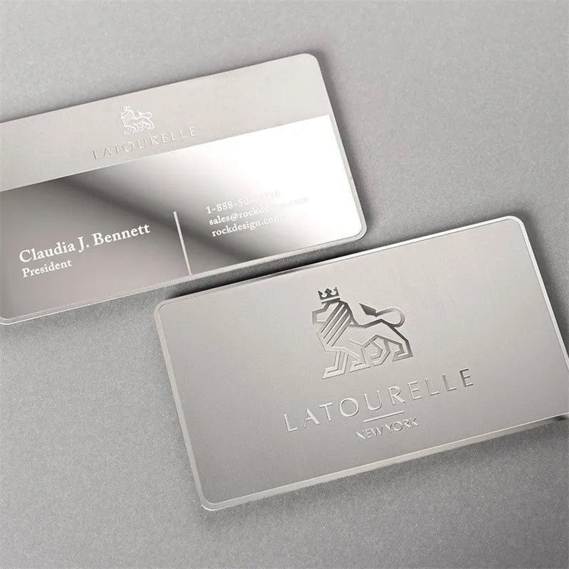 Two silver-tone metal business cards with fine engraved-style artwork on a pale background