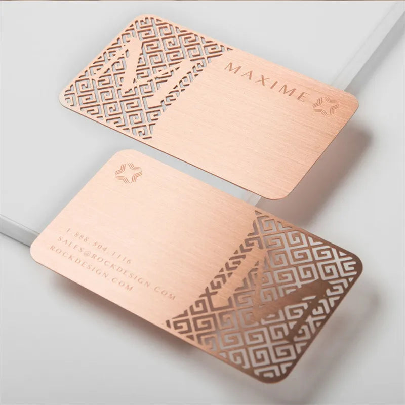 Two rose-gold-tone metal cards with decorative cutout borders on a pale surface