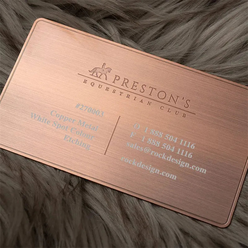 Copper-tone metal business card with fine engraved-style lettering displayed on wood