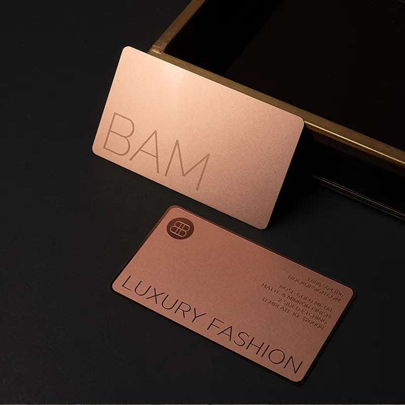 Two copper-tone metal business cards with minimalist lettering against a dark background