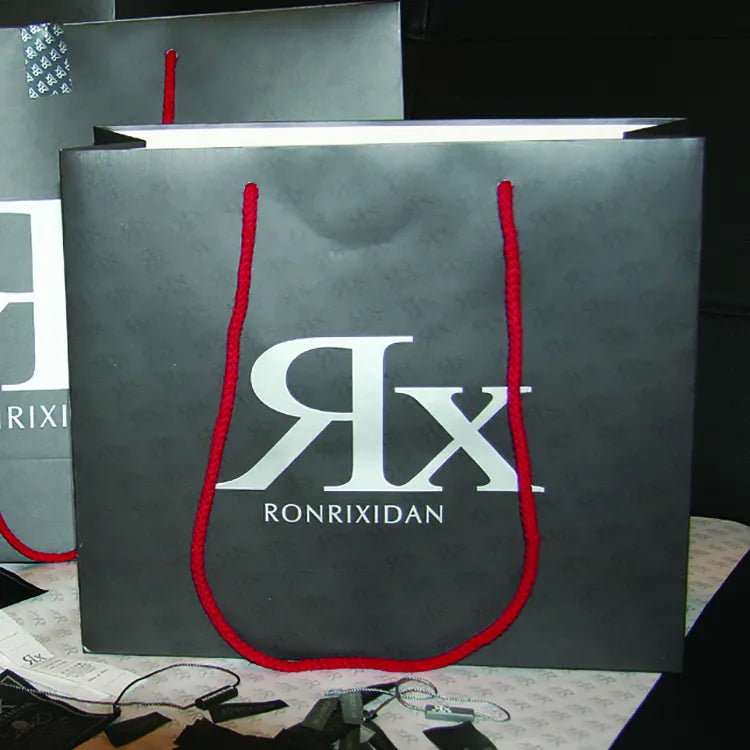 Gray paper shopping bag with red cord handles and a large contrasting printed logo