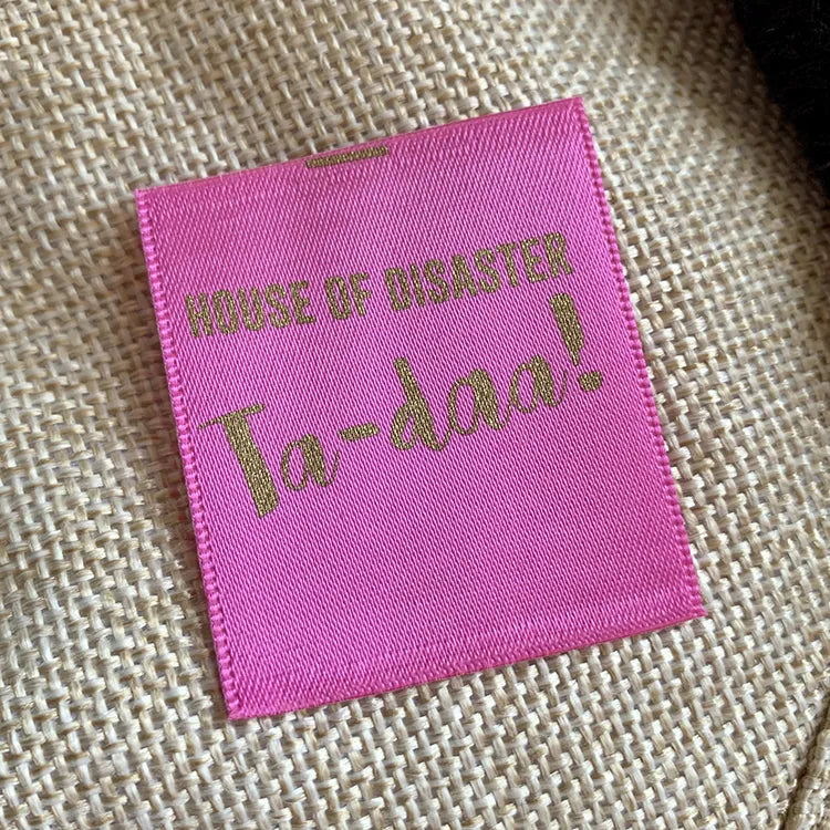 Bright pink rectangular clothing label with metallic gold-colored lettering