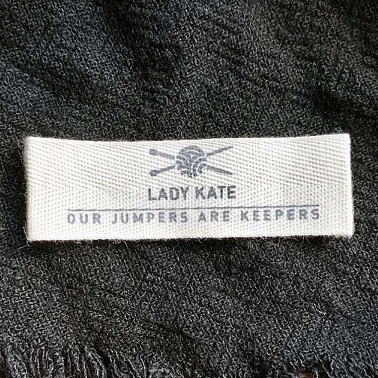 Small light-colored fabric label with dark text and a scissors emblem on black cloth