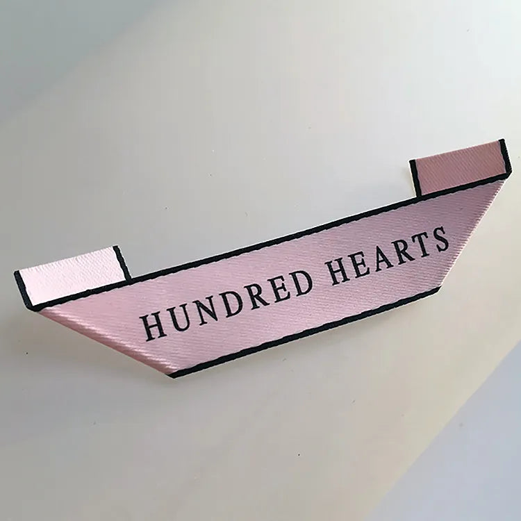 Pink folded clothing brand label with black HUNDRED HEARTS lettering and a border