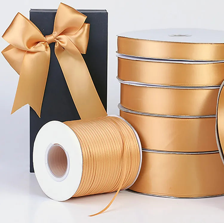 Gold satin ribbon rolls in assorted widths beside a tall black gift box tied with a satin bow