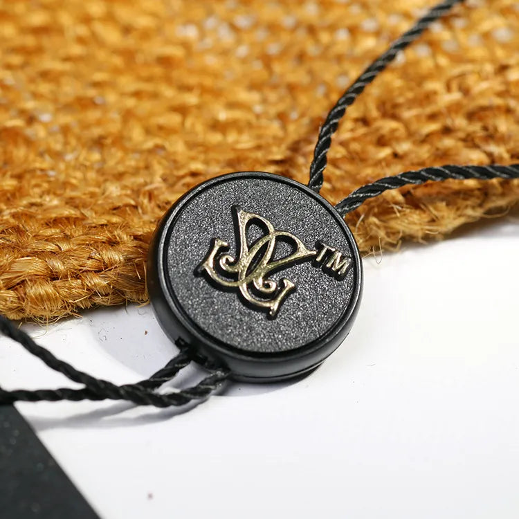 Round black plastic button tag with gold-tone monogram emblem and black twisted cords