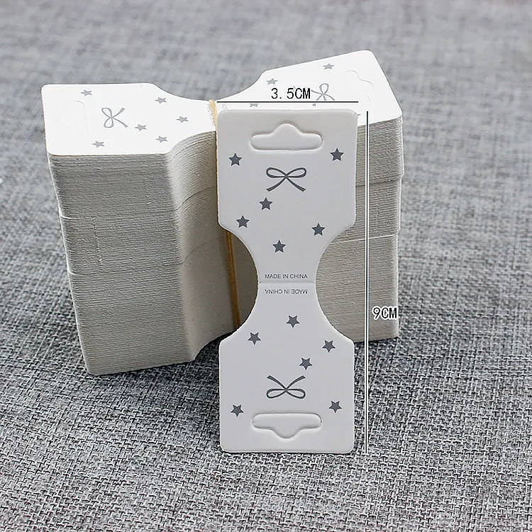 Stack of white foldover jewelry display cards (3.5cm x 9cm) with die-cut euro hanging slot, printed star and bow motif, and center fold line