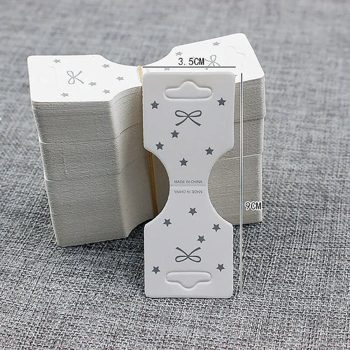 Stack of white foldover jewelry display cards (3.5cm x 9cm) with die-cut euro hanging slot, printed star and bow motif, and center fold line