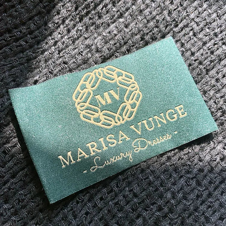 Teal woven clothing label with gold Marisa Vunge logo and lettering