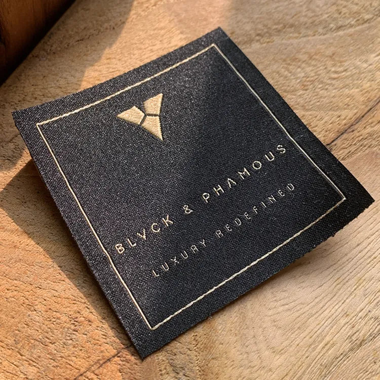 Black woven clothing label with gold border and emblem, featuring “Black Is Famous” text