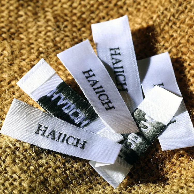 White woven clothing labels with dark green lettering, arranged in a fan shape on mustard yellow knitted fabric