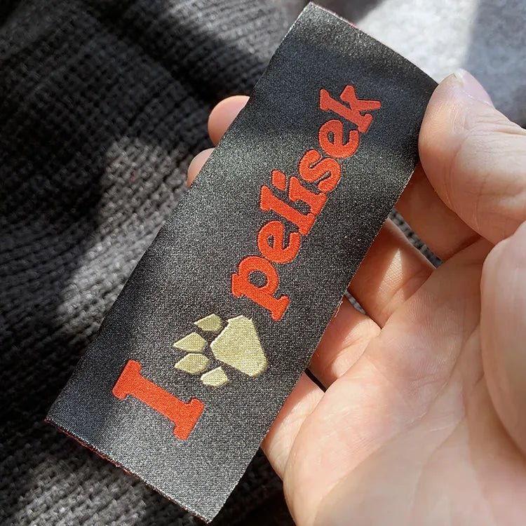 Hand holding a black woven label with red lettering and a pale gold paw print